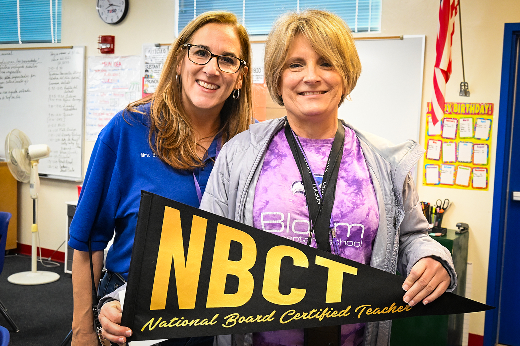 A woman in a blue shirt and glasses smiles with a woman in a purple shirt holding an NBCT pennant