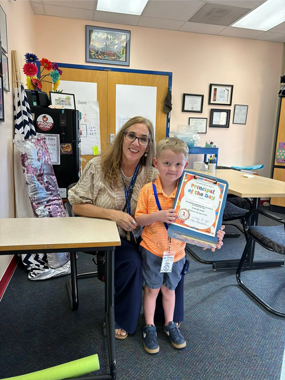 A woman kneels next to a small boy holding a Principal of the Day certificate